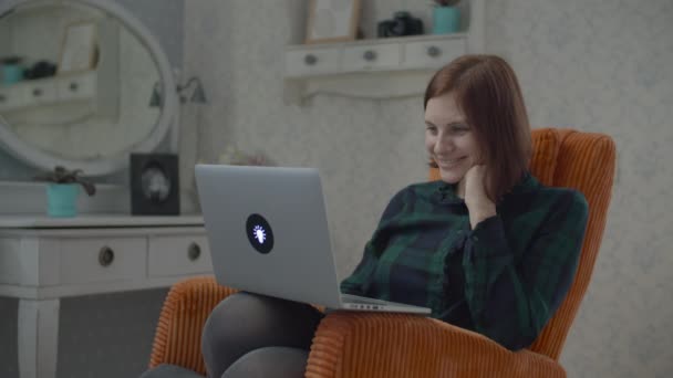 Jovem morena adulta assistindo conteúdo on-line no laptop sentado em poltrona laranja acolhedora no quarto. Tela Gadget piscando para o rosto da mulher . — Vídeo de Stock