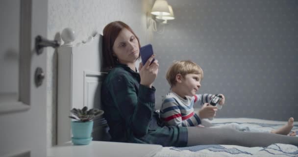 Jovem mãe com smartphone nas mãos e filho jogando videogames sentados na cama em casa. Passatempo da noite em família. Menino ganhando em videogame e levantando as mãos — Vídeo de Stock