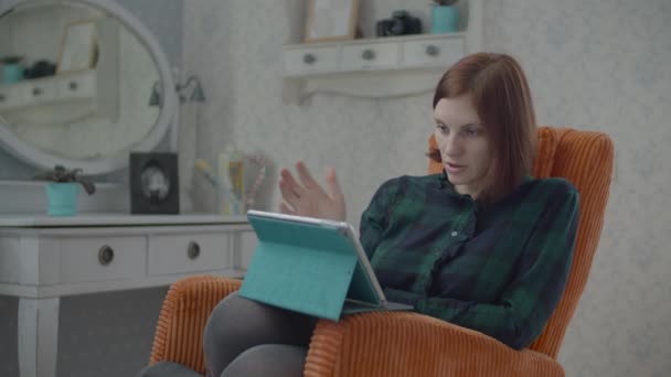 Young female brunette adult talking by tablet computer on her knees. Woman sitting in cosy orange armchair in bedroom. — Stock Video