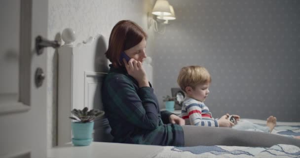 Genç bir anne akıllı telefon ve oğluyla konuşur video oyunları oynar, evde yatağında cips yer. Aile akşamı eğlencesi. — Stok video