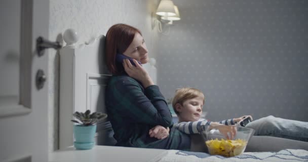 Jovem mãe falando por smartphone e filho jogando videogames, comendo fichas sentadas na cama em casa. Passatempo da noite familiar . — Vídeo de Stock
