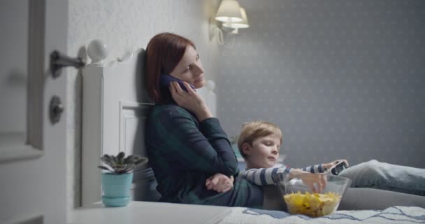 Ung mor talar via smartphone och son spelar tv-spel, äter chips sitter på sängen hemma. Familjekväll. — Stockvideo