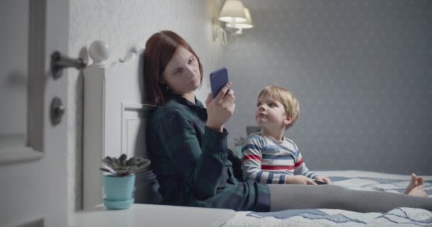 Jeune mère avec smartphone dans les mains et fils jouant à des jeux vidéo assis sur le lit à la maison. Passe-temps familial. Garçon dérangeant mère . — Video