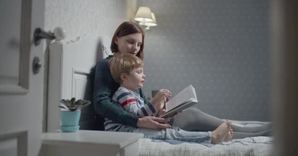 Mutter und Vorschulsohn lesen Papierbuch auf dem Bett zu Hause. Kind liest Buch mit Mama vor. glückliche Familienzeit. — Stockvideo