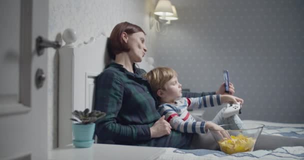 Joven madre grabando video selfie por teléfono inteligente e hijo jugando videojuegos sentados en la cama en casa. Pasatiempo familiar. Chico ganando en el videojuego y levantando las manos — Vídeos de Stock