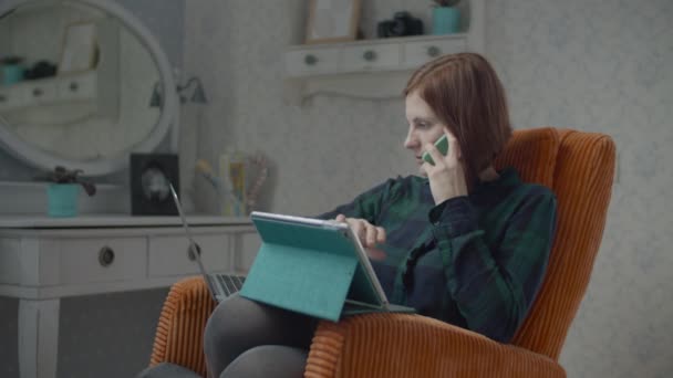 Fiatal nő barna felnőtt beszél okostelefonon tabletta számítógép és laptop a térdét. Nő ül kényelmes narancssárga fotelben a hálószobában. — Stock videók