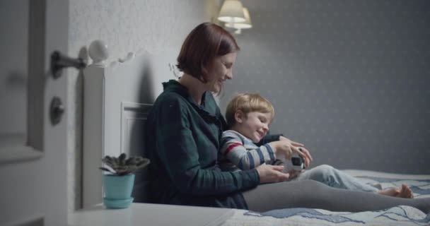 Feliz madre e hijo jugando videojuegos en la cama en casa. Niño sosteniendo el controlador inalámbrico en las manos, mujer enseñando a jugar. Familia divirtiéndose . — Vídeo de stock
