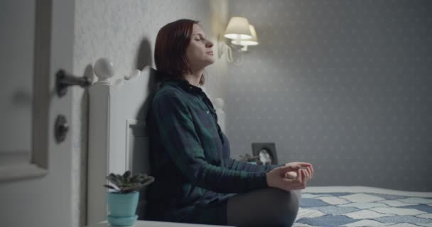 Молода жінка сидить в позі лотоса медитуючи з мобільним додатком на ліжку. Жінка відчуває спокій, роздумуючи ввечері . — стокове відео