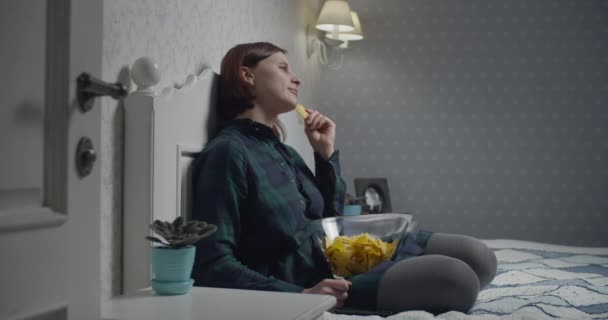Junge weibliche Erwachsene genießen fernsehen mit Chips Schüssel auf dem Bett. Frau schaltet Fernseher abends per Fernbedienung aus — Stockvideo