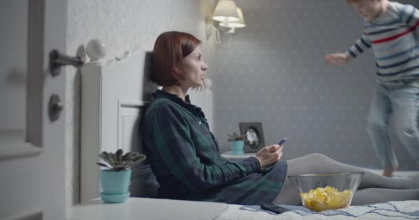 Mutter und Sohn amüsieren sich beim Fernsehen zu Hause auf dem Bett. Kind springt auf Bett, während Mama fernsieht. — Stockvideo