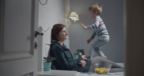 Mutter und Sohn amüsieren sich beim Fernsehen zu Hause auf dem Bett. Kind springt auf Bett, während Mama fernsieht. — Stockvideo