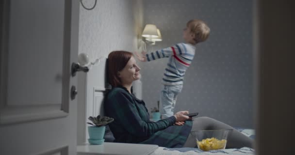 Mutter und Sohn amüsieren sich beim Fernsehen zu Hause auf dem Bett. Kind springt auf Bett, während Mama fernsieht. — Stockvideo