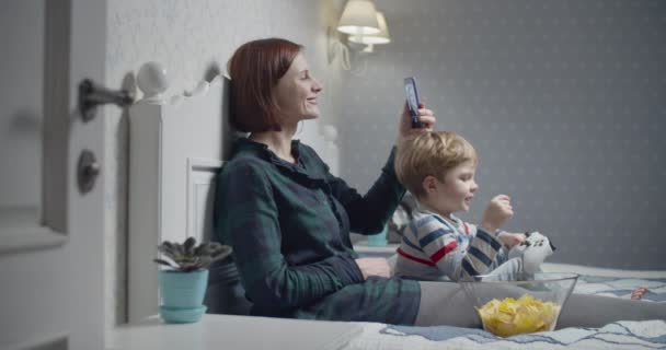 Mladá matka nahrává selfie video chytrým telefonem a syn hraje videohry sedící doma v posteli. Rodinná večerní zábava. Chlapec vítězí ve videohře a zvedá ruce — Stock video