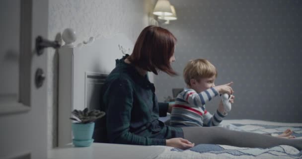 Счастливая мать и сын играют в видеоигры на кровати дома. Мальчик держит беспроводной контроллер в руках, женщина учит играть. Семья веселится . — стоковое видео