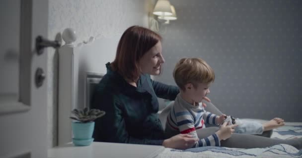 Šťastná matka a syn hrají videohry doma v posteli. Chlapec drží bezdrátový ovladač v rukou, žena učí hrát. Rodina se baví. — Stock video