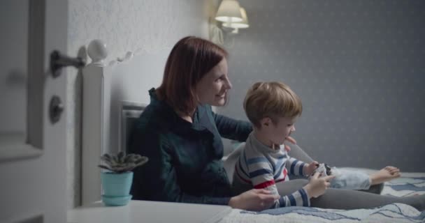 Happy mother and son playing video games on bed at home. Boy holding wireless controller in hands, woman teaching how to play. Family having fun. — 비디오