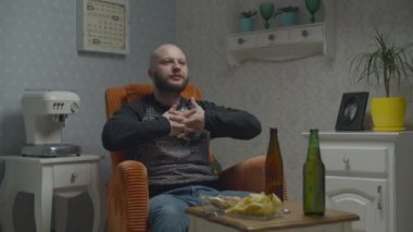 Kel sakallı adam ısınıyor. Bira ve cipsli televizyon izlerken rahat koltukta oturuyor.. 