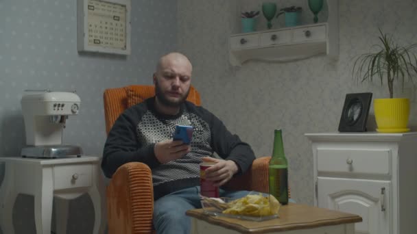 Homme chauve barbu surfer en ligne sur smartphone tout en regardant la télévision et boire de la bière avec du poisson et des frites assis dans un fauteuil confortable . — Video