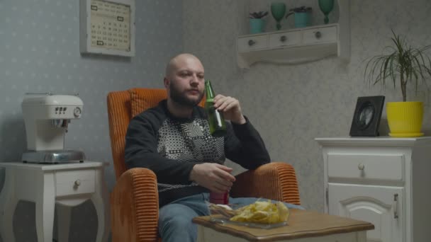 Łysy, smutny brodaty facet oglądający telewizję i pijący piwo z rybą i frytkami. mężczyzna otwarcie butelki piwa siedzi w przytulny fotel. — Wideo stockowe