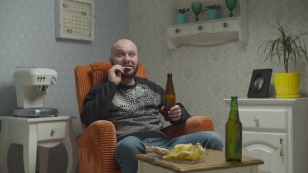 Skæg mand nervøst ser tv og drikker øl med fisk og chips. Mand fejrer sejr på tv-skærmen sidder i hyggelig lænestol. Triumfalt hænder op . . – Stock-video