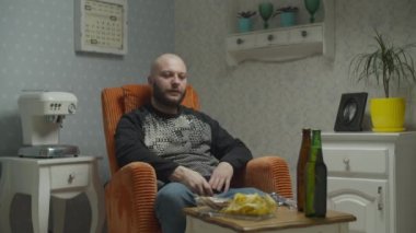 Kel sakallı adam televizyon izliyor ve balık ve patates kızartmasıyla bira içiyor. Erkek televizyonu uzaktan kumandayla açıyor ve bira şişesini açıyor.. 
