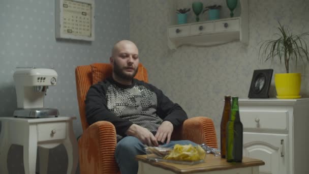 En skallig skäggig man som tittar på TV och dricker öl med fish and chips. Man slår på Tv med fjärrkontroll och öppna öl flaska. — Stockvideo