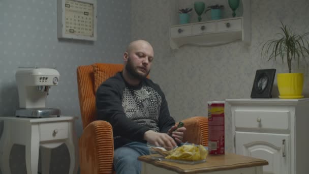 Bald sad bearded man watching TV and drinking beer with fish and chips. Male opening beer bottle sitting in cozy armchair. — Stock Video