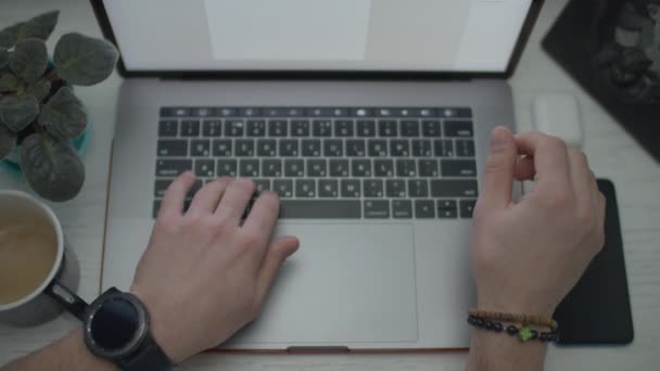 Мужские руки с часами, печатающими на ноутбуке и пьющими кофе. Руки работают на клавиатуре. Вид сверху ноутбука, смартфона, наушников и чашки кофе на столе . — стоковое видео