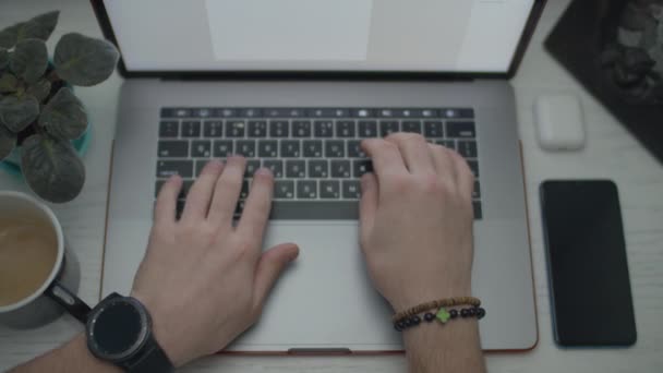 ノートパソコンに文字盤を持った腕時計を手にする。キーボードを操作する手。ノートパソコン、スマートフォン、イヤホン、コーヒーカップのトップビュー. — ストック動画
