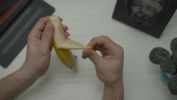 Le mani di uomo sbucciano una banana sopra la scrivania con computer di targa. Peeling banana vista dall'alto . — Video Stock