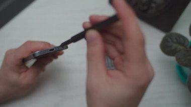 Beyaz ahşap masadaki küçük portatif tornavidayla akıllı telefonu tamir eden bir adam. Adam hücredeki vidayı gevşetip sıkıyor.. 