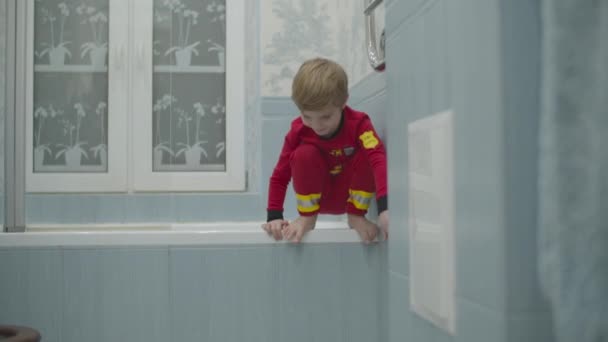 Niño rubio preescolar en pijama de bombero divirtiéndose en el piso del baño. Niño jugando alrededor saltando de la bañera al suelo . — Vídeos de Stock