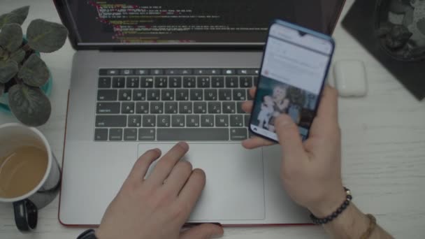 Mans mani navigare online tramite cellulare durante la codifica sul computer portatile. L'uomo inizia ad inserire codice sulla tastiera. Vista dall'alto di laptop, smartphone, auricolari e tazza di caffè sulla scrivania . — Video Stock