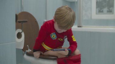 Sarı kreş çocuğu itfaiyeci pijamalarıyla tuvalette oturmuş mavi banyoda akıllı telefon izliyor.. 