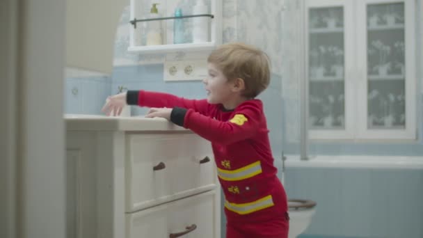 Niño rubio preescolar en pijama de bombero corriendo al lavabo, tomando cepillo de dientes y empezando a cepillarse los dientes en el baño . — Vídeos de Stock