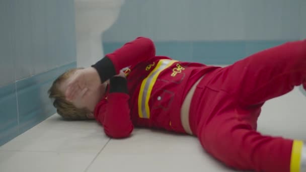 Blond kleuter in brandweerpyjama die plezier heeft op de badkamervloer. Kind rotzooit rond op de witte vloer. — Stockvideo