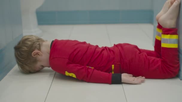 Blond förskola pojke i brandman pyjamas har roligt på badrumsgolvet. Ungen lurar runt på vitt golv. — Stockvideo