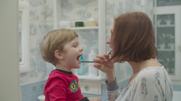 Jonge moeder helpt kleuter in rode pyjama met tanden poetsen met tandzijde. Leuke familie bad routine. Moeder en zoon lachen in de badkamer. — Stockvideo