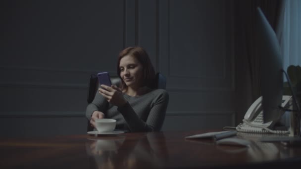 Junge Geschäftsfrau, die nachts im Büro arbeitet. Frau surft online mit Smartphone und trinkt Kaffee im gemütlichen Büroledersessel. — Stockvideo