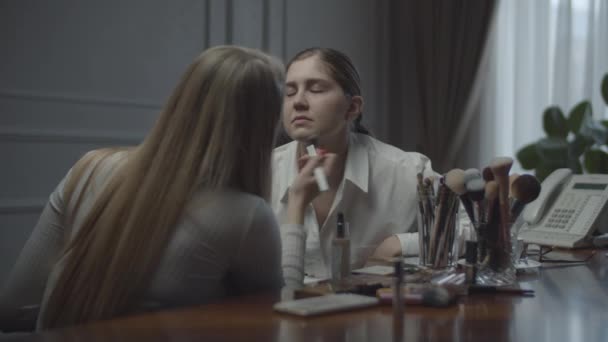 Deux collègues féminines postulant se font maquiller au bureau. Visagiste s'applique maquillage pour femme d'affaires sur le lieu de travail. Beaucoup de cosmétiques et brosses sur la table . — Video