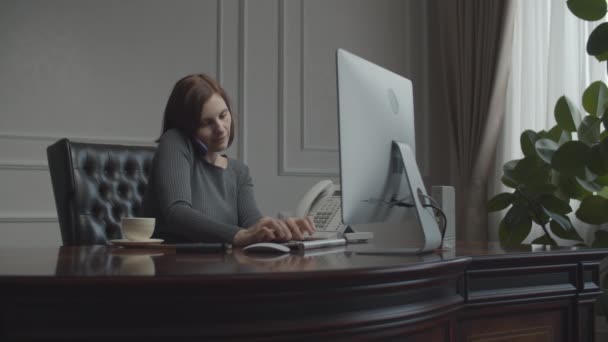 Jeune femme d'affaires parlant sur smartphone. Brunette travaillant au bureau avec moniteur assis dans le fauteuil dans le bureau d'affaires . — Video