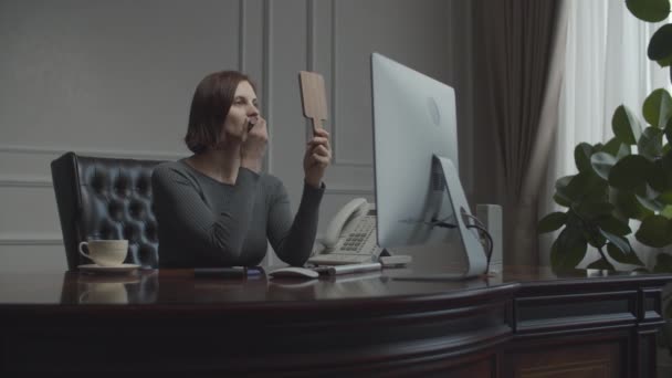 Young businesswoman applying make up looking in mirror at working place. Brunette in business office talking on smartphone. Desk with monitor. — 비디오
