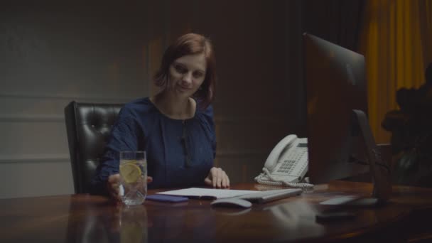 Молодая деловая женщина, работающая в ночном офисе. Женщина кладет в стакан воды лимонный мир и пьет его. Работающая женщина за столом с монитором . — стоковое видео