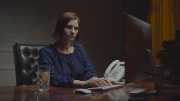 Молода бізнес-леді працює в нічному офісі. Жінка друкує на клавіатурі і п'є воду з лимоном. Працююча жінка за столом з монітором. Вид спереду . — стокове відео