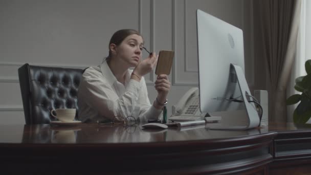 Jonge zakenvrouw die make-up aanbrengt, kijkt in de spiegel naar het werk. Vrouw aan het bureau met monitor in het kantoor. — Stockvideo