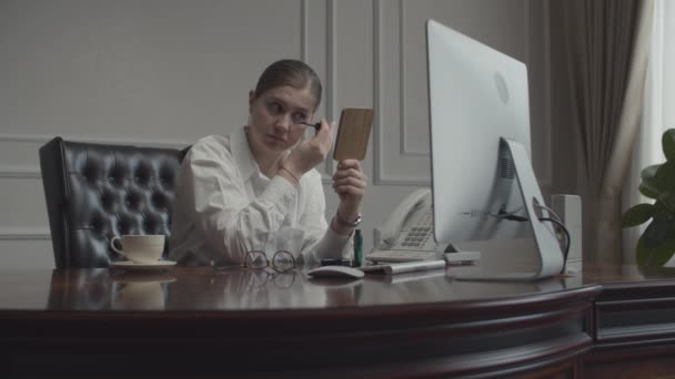 Jovem empresária aplicando maquiagem olhando no espelho no local de trabalho. Feminino sentado na mesa com monitor no escritório de negócios . — Vídeo de Stock