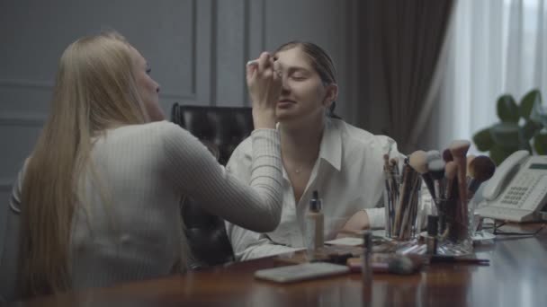 Duas colegas a candidatar-se compõem o cargo. Visagiste aplica compõem a mulher de negócios no lugar de trabalho. Lotes de cosméticos e escovas na mesa . — Vídeo de Stock