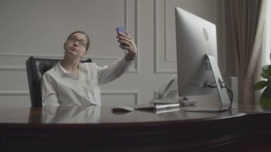 İş yerindeki koltukta monitörle çalışan genç bir iş kadını. Özçekim yapmak için akıllı telefondan dikkati dağılmış bir kadın.