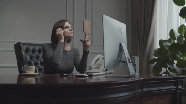 Young businesswoman applying make up looking in mirror at working place. Brunette in business office talking on smartphone. Desk with monitor. — Stock Video