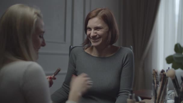 Two female friends colleagues applying make up in office and laughing. Woman applies make up to friend on working place. Lots of cosmetics and brushes on table. — Stock Video